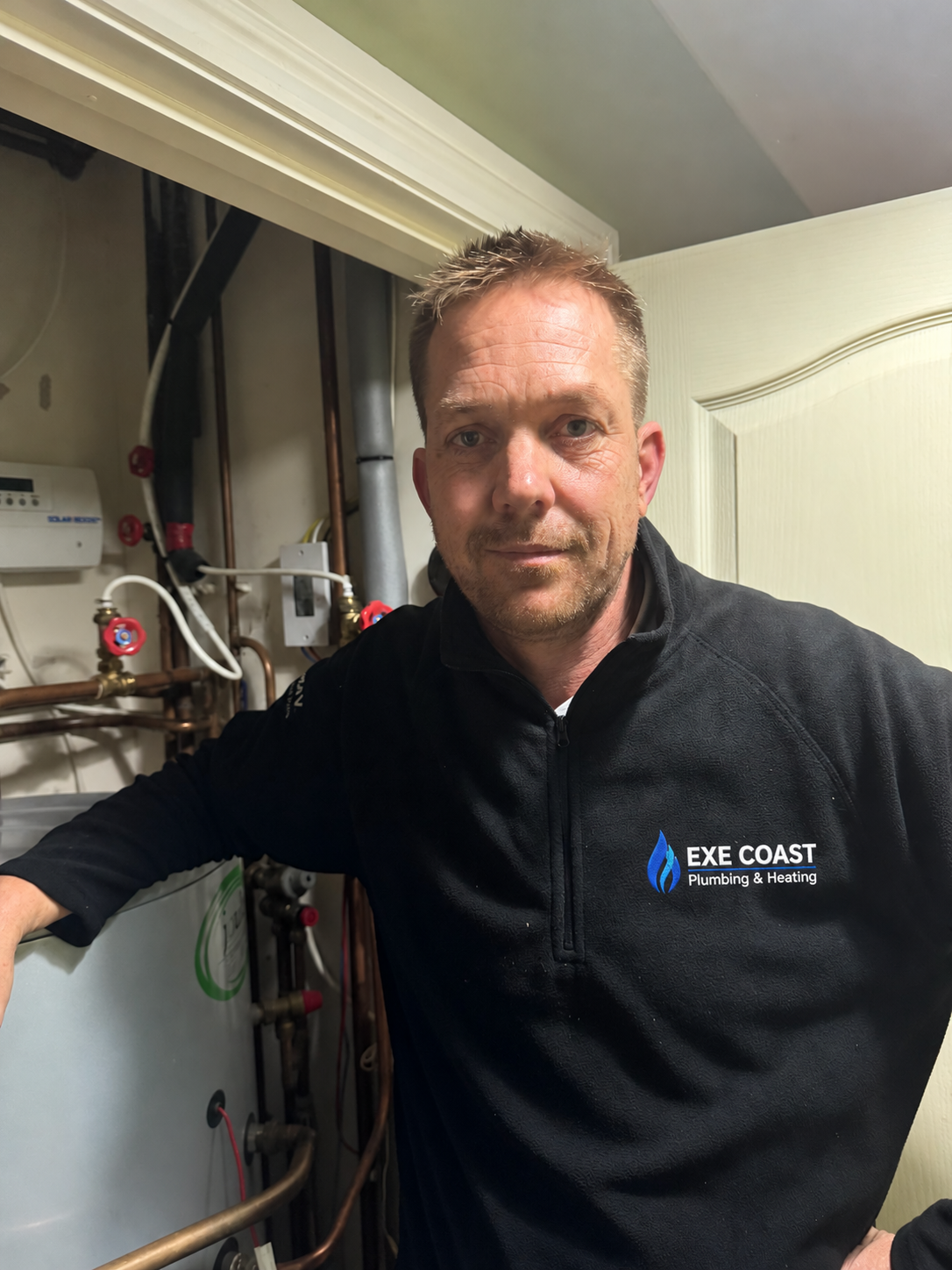 Grant beside plumbing pipework, wearing an Exe Coast fleece